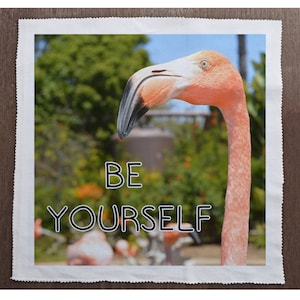 May include: A pink flamingo with a black beak and a long neck is the focal point of this image. The words "BE YOURSELF" are written in black outlined letters. The background features green foliage and a blue sky.