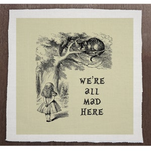 May include: A black and white illustration of Alice from Alice in Wonderland standing in a forest with a Cheshire Cat sleeping in a tree above her. The text "We're all mad here" is printed below the image.