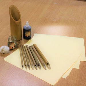May include: A set of 10 bamboo calligraphy pens with a bamboo pen holder, ink bottle, and ink jar. The pens are arranged on a stack of yellow paper.