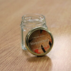 May include: A small, clear glass jar with a gold lid. The lid has a label that says "Calligraphy Supplies Maryam Ovais" and a website address.