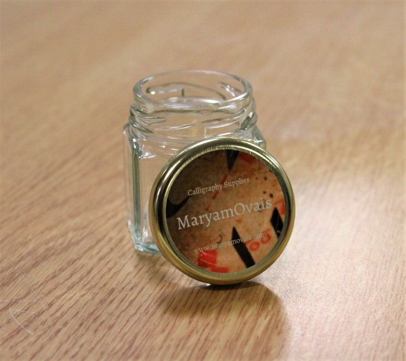 May include: A small, clear glass jar with a gold lid. The lid has a label that says "Calligraphy Supplies Maryam Ovais" and a website address.