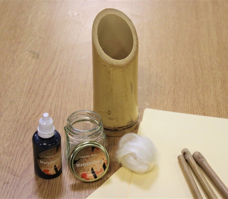 May include: Calligraphy supplies including a bamboo pen holder, a glass jar with a gold lid, a bottle of black ink, a small ball of white cotton, and three wooden calligraphy pens on a yellow surface.