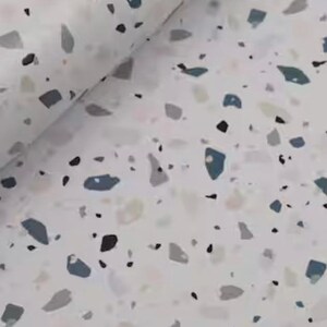 May include: A roll of white paper with a terrazzo pattern. The design features irregular shapes in shades of gray, blue, and black scattered across the surface. The paper is slightly rolled, showing the pattern's repeating nature.