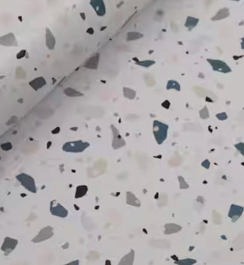 May include: A roll of white paper with a terrazzo pattern. The design features irregular shapes in shades of gray, blue, and black scattered across the surface. The paper is slightly rolled, showing the pattern's repeating nature.