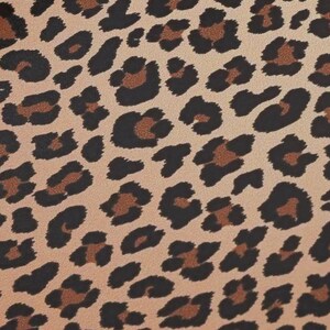 May include: Close-up of a tan fabric with a leopard print pattern. The pattern features black and brown spots of varying sizes and shapes, creating a textured, animal-inspired design. The background is a light tan color.