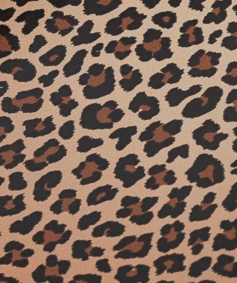 May include: Close-up of a tan fabric with a leopard print pattern. The pattern features black and brown spots of varying sizes and shapes, creating a textured, animal-inspired design. The background is a light tan color.