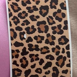 May include: A rectangular sticker with a leopard print design. The sticker features a tan background with dark brown and black spots. The sticker has a white border and a scalloped top edge. A pink surface is visible on the left side of the sticker.