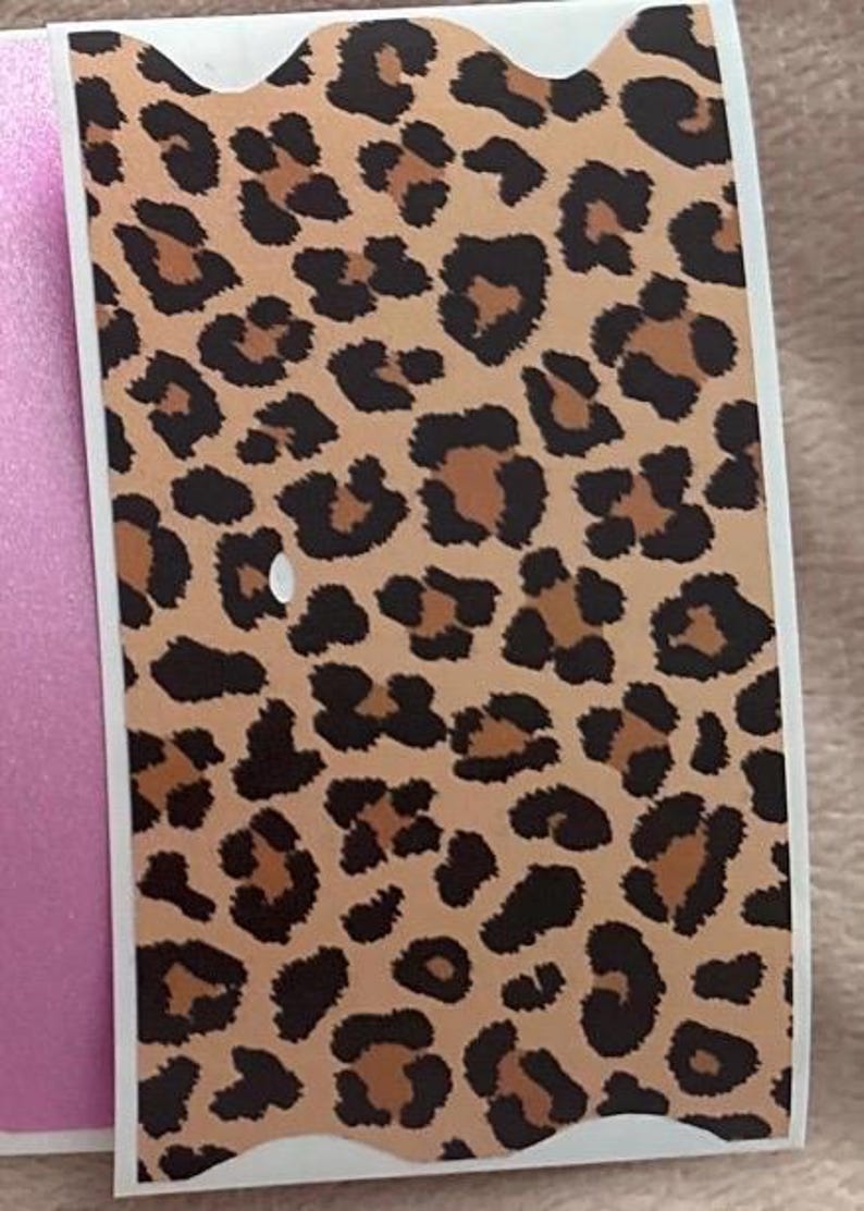 May include: A rectangular sticker with a leopard print design. The sticker features a tan background with dark brown and black spots. The sticker has a white border and a scalloped top edge. A pink surface is visible on the left side of the sticker.