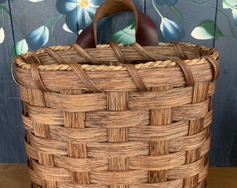Può includere: Un cesto intrecciato di forma rettangolare con un manico in pelle marrone. Il cesto è realizzato con strisce intrecciate di materiale marrone chiaro e scuro. Lo sfondo presenta un motivo floreale con fiori blu.