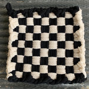 May include: A square, woven potholder with a black and white checkerboard pattern. The edges are trimmed with a black, braided border and a small loop for hanging. The potholder is made of a natural fiber.