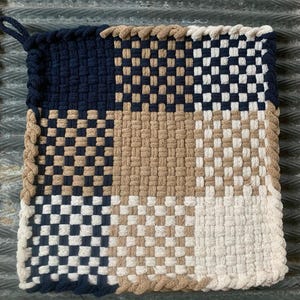 May include: A square, woven pot holder with a loop for hanging. The design features a checkerboard pattern in navy blue and white, with sections of tan and off-white. The edges are trimmed with a braided border.