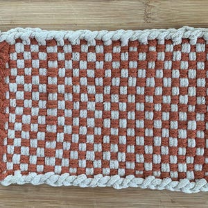 May include: A rectangular woven potholder featuring a checkered pattern of terracotta orange and off-white fabric strips. The edges are finished with a braided border in alternating colors, and a white plastic ring is attached to one corner.
