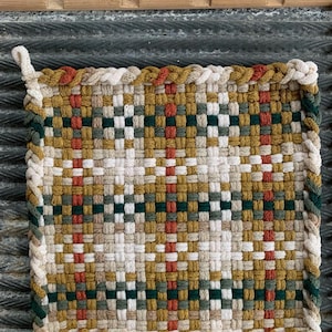 May include: A square, woven hot pad with a loop for hanging. The pad features a geometric pattern in shades of green, gold, red, and cream. The edges are finished with a twisted rope trim in complementary colors. The hot pad is designed for kitchen use.