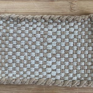 May include: A rectangular woven potholder featuring a checkered pattern in alternating light beige and tan colors. The edges are finished with a thick, braided border in matching tones, and a small white circular loop is attached to the top corner.