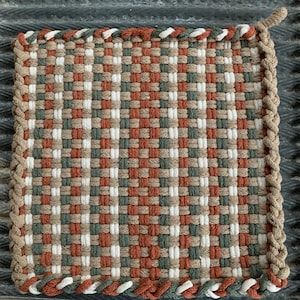 May include: Square woven hot pad with a checkerboard pattern in shades of white, brown, green, and rust. The edges are finished with a braided border in a neutral beige color. A small loop is attached to one corner.