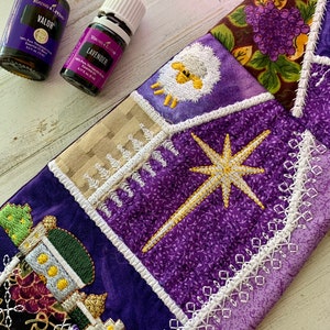 Può includere: Tessuto viola e bianco con disegni ricamati, tra cui una stella, un agnello e una vite. In primo piano ci sono due piccole bottiglie di olio essenziale, etichettate "Valor" e "Lavender".