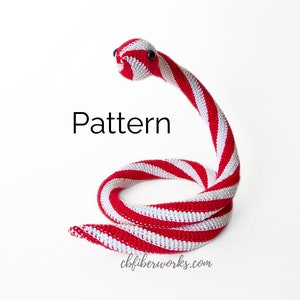 May include: A red and white striped crocheted snake toy. The snake is curled up in a circle with its head raised. The text "Pattern" is written in black above the snake. The text "cbfiberworks.com" is written in black below the snake.