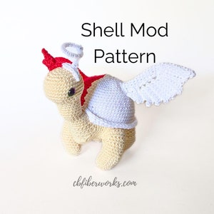 Puede incluir: Una tortuga de ganchillo con cuerpo de color beige claro, un tocado rojo y un halo blanco. Tiene un caparazón azul claro y alas blancas. El texto "Shell Mod Pattern" está encima de la tortuga. El sitio web cbfiberworks.com está en la parte inferior.
