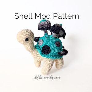 May include: A crocheted turtle amigurumi toy with a teal shell and black and grey polka dots. The turtle has a beige body and three grey and black mushroom-shaped decorations on its back. The text "Shell Mod Pattern" is above the toy and "cbfibworks.com" is below the toy.