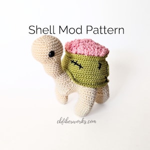 Op de afbeelding: Een gehaakt amigurumi-speelgoed in de vorm van een schildpad met een roze en groene schild. De schild heeft een gestikt gezicht met zwarte ogen en een gestikte mond. De tekst "Shell Mod Pattern" staat boven het speelgoed. De tekst "cbfiberworks.com" staat onder het speelgoed.