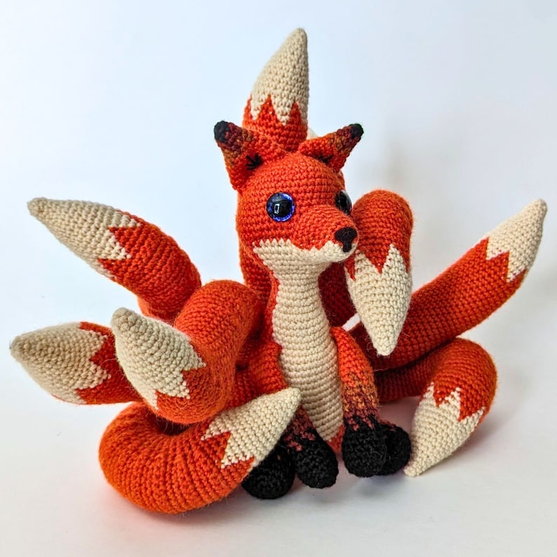 May include: A crocheted fox with eight tails, in shades of orange, cream, and black. The fox has large blue eyes and is sitting upright. The tails are fanned out, and the fox has a friendly expression. The craft is handmade.