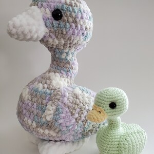 May include: Two crocheted duck plushies. The larger duck is made with a multi-colored yarn in shades of white, gray, lavender, and blue. The smaller duck is a solid light green color.