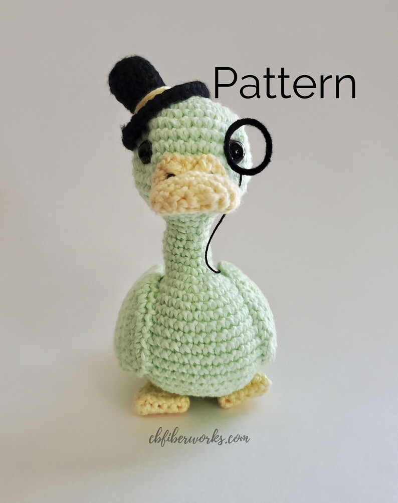 May include: A green crocheted duck wearing a black top hat and a monocle. The duck has a yellow beak and feet. The text "Pattern" is visible in the upper right corner of the image.