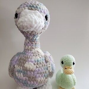 May include: Two crocheted duck plushies. The larger duck is made with a multi-colored yarn in shades of white, gray, blue, and purple. The smaller duck is a solid light green color. Both ducks have black eyes and yellow beaks.