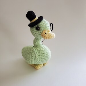 May include: A green crocheted duck wearing a black top hat and glasses. The duck has yellow feet and a yellow beak.