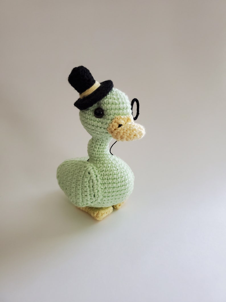May include: A green crocheted duck wearing a black top hat and glasses. The duck has yellow feet and a yellow beak.