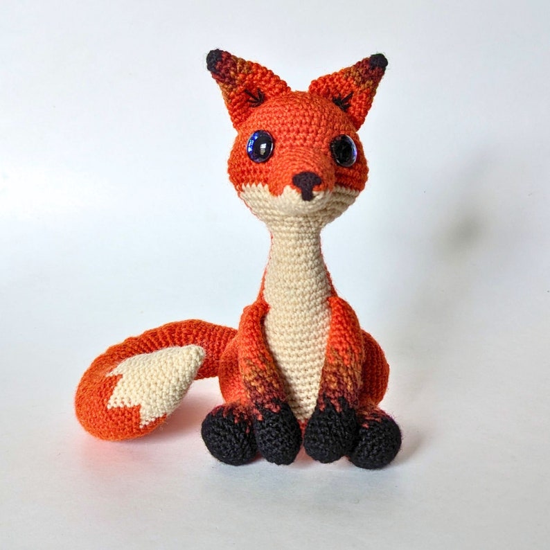 May include: A handmade crochet fox toy with orange, white, and black details. The fox has large, blue eyes and a long, curved tail. The fox is sitting upright on a white surface.