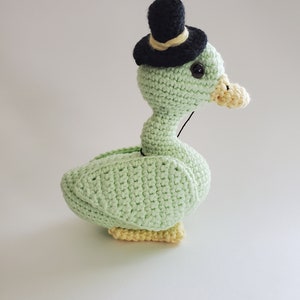 May include: A green crocheted duck wearing a black top hat with a yellow band. The duck has black eyes and a yellow beak.