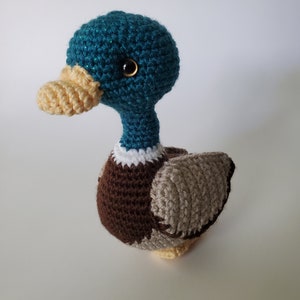 May include: A teal blue crocheted duck with brown and beige feathers. The duck has a yellow beak and black eyes.