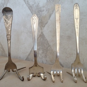 May include: Four silver forks have been bent into a tripod shape to create a display stand for small items. The forks have decorative designs on the handles.