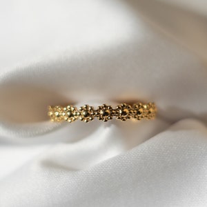 Könnte beinhalten: Ein goldfarbener Ring mit floralem Design. Der Ring besteht aus kleinen, miteinander verbundenen blütenförmigen Elementen. Der Ring ist vor einem weißen, seidigen Hintergrund platziert.