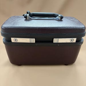 May include: A rectangular dark brown hard-shell travel case with a textured finish. The case features a black plastic handle with chrome-coloured metal accents, a black trim, and two silver-toned metal locking mechanisms on the front panel.