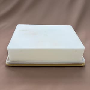 May include: A rectangular, off-white plastic butter dish with a matching lid. The dish sits on a yellow-toned base. The lid has a circular indentation on top. The butter dish is a kitchen storage item.