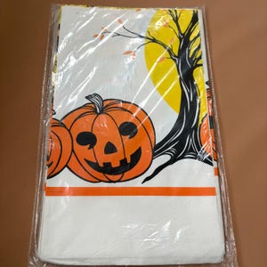 Puede incluir: Un mantel de Halloween con calabazas naranjas, un árbol negro y una luna amarilla sobre un fondo blanco. Una franja naranja recorre la parte inferior. El mantel está sellado en plástico.