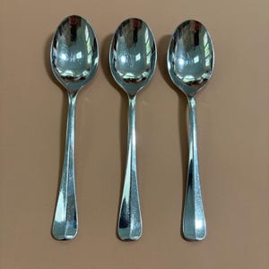 May include: Three identical silver-toned metal spoons arranged vertically side by side. Each spoon features a smooth, polished finish with a reflective surface and a simple, tapered handle design that widens slightly toward the base of the stem.