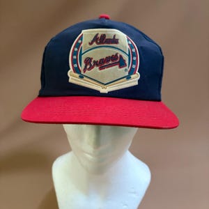 May include: A navy blue baseball cap with a red brim. The front features a cream-colored patch with the text Atlanta Braves in red and blue script, surrounded by a circular border with small baseball icons and two crossed baseball bats.