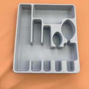 May include: A light gray plastic utensil organizer with multiple compartments. The organizer has sections for forks, knives, spoons, and other kitchen tools. The organizer is rectangular and has rounded edges.