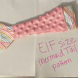 May include: Pink and rainbow fabric mermaid tail pattern for an elf size. The tail is made of a soft, textured fabric with a rainbow pattern on the top and a pink, textured fabric on the bottom.