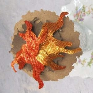 May include: A paper craft decoration featuring a stylized flame design with gold and orange metallic paper. The flame is layered on a brown paper circle.