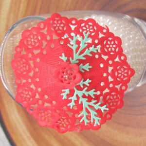 May include: Red lace doily with a green snowflake design. The doily is on top of a glass jar filled with white beads.