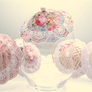 May include: A set of six floral paper doilies with lace trim, each with a small white paper umbrella on top. The doilies are arranged in a glass vase.