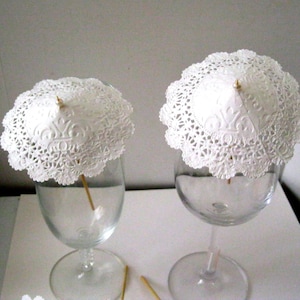 May include: Two white paper doilies with scalloped edges are shaped into small umbrellas and placed on top of clear glass wine glasses. The umbrellas are attached to wooden skewers.