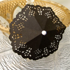May include: A black paper parasol with a scalloped, lace-like edge. The parasol is open, revealing its interior structure and a small, white and blue decorative element at the top. The parasol is set against a blurred background.