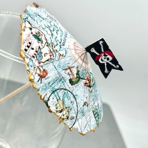May include: A cocktail umbrella with a nautical map design and a small black flag featuring a skull and crossbones. The umbrella has a light blue and beige color scheme, with a gold-colored edge.