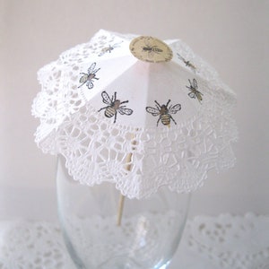 May include: A white paper cocktail umbrella with a lace trim and bee designs. The umbrella is on a wooden stick and is in a clear glass.