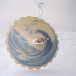 May include: A round, scalloped paper card with a blue watercolor wave design and a small seashell on top. The card is beige with a light brown border.